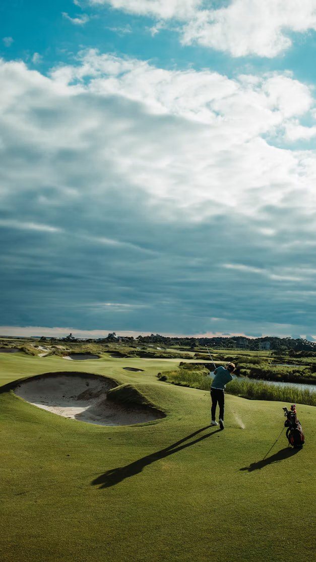 A golfer walking a fairway beside a bunker under a big sky