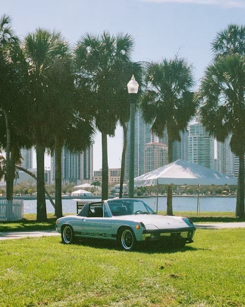 A vintage car parked on a lawn under palm trees with a city skyline behind